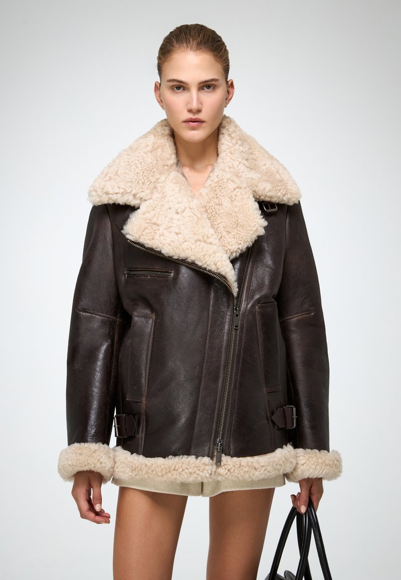 Giacca in shearling marrone con un ampio colletto beige, dotata di chiusura con zip, tasche laterali e dettagli con fibbie all'orlo e alle maniche.
