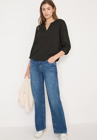 Schwarze Bluse mit langen Ärmeln und V-Ausschnitt, kombiniert mit weitem blauen Jeansstoff und weißen Turnschuhen. Struktur des Stoffes und Denims sichtbar.