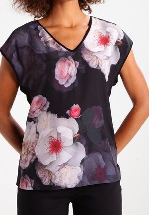 Femme portant un top noir à col en V avec un grand imprimé floral rose et blanc, manches courtes et coupe décontractée.