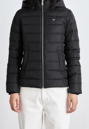 Personne portant une veste matelassée noire à capuche avec une fermeture éclair à l'avant et un pantalon blanc, debout devant un fond uni.