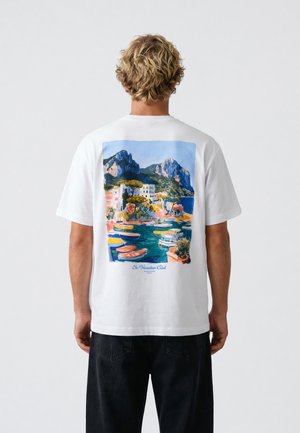 Mand med blond krøllet hår iført hvid t-shirt med farverigt kystlandskabsmaleri og både på ryggen, sorte bukser, stående.