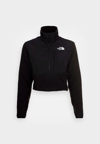 Chaqueta corta negra con un alto cuello, que presenta un cuerpo de forro polar y una sección superior lisa. Incluye un bolsillo con cremallera en la parte frontal y un logo.