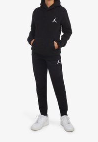 Felpa nera con cappuccio e tasca frontale, logo bianco sul petto, abbinata a pantaloni sportivi neri e sneakers bianche.