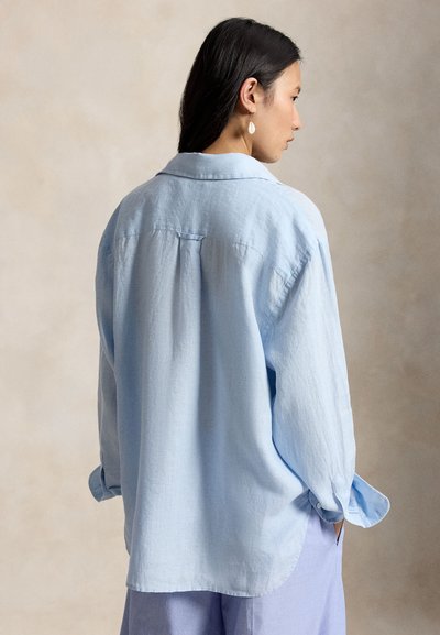 Žena s dlhými tmavými vlasmi, oblečená v voľnej svetlomodrej košeli a levanduľových nohaviciach, stojaca bokom s rukami v džepoch na neutrálnom pozadí.