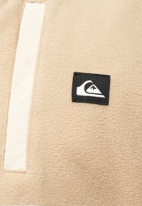 Μπεζ ύφασμα φλις με ραμμένο μαύρο λεκέ που διαθέτει ένα στυλιζαρι σμένο λευκό κύμα και λογότυπο βουνού δίπλα σε μια κάθετη λευκή λωρίδα.