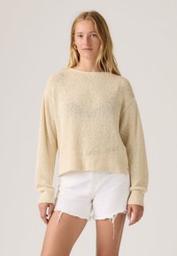 Γυναίκα με μακριά ξανθά μαλλιά που φοράει μπεζ πλεκτό πουλόβερ και λευκά σκισμένα σορτς, στέκεται μπροστά σε απλό φόντο.