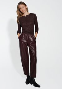 Donna che indossa un maglione button-up marrone scuro, pantaloni larghi in ecopelle borgogna e stivaletti neri, con le mani in tasca.