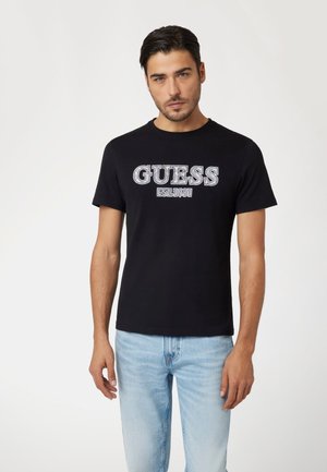 Juodas medvilnės marškinėliai su baltu „GUESS EST.1981“ grafiku drąsiu šriftu. Klasikinis apvalus iškirpimas ir trumpomis rankovėmis, derinami su šviesiai mėlynomis džinsinėmis kelnėmis.