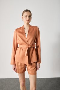Peach-colored blazer and shorts set, made of smooth fabric, featuring a waist tie, notched collar, and long sleeves. Minimalist design.