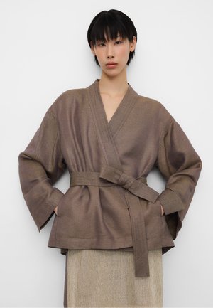 Vrouw met kort zwart haar, gekleed in een losse bruine wikkeljas met brede mouwen en een stoffen riem die in de taille is gestrikt.