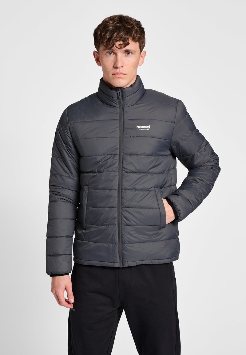 Jonge man draagt een donkergrijze doorgestikte jas met ritszakken en zwarte broek, staand met één hand in de zak tegen een witte achtergrond.