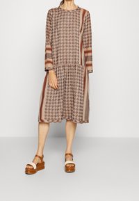 Hnědé vzorované šaty s dlouhými rukávy, kulatým výstřihem a nařaseným pasem. Slučující se s hnědými platformovými sandály.