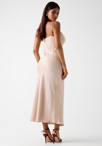 Długa, dopasowana suknia z miękkiego beżowego materiału z subtelnym wzorem kropek, na cienkich ramiączkach, o gładkiej fakturze. Do tego bordowe sandały na wysokim obcasie.