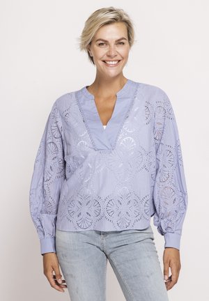 Lächelnde Frau mit kurzen blonden Haaren trägt eine hellviolette Bluse mit Spitzenmuster, langen Ballonärmeln und hellblauen Jeans.