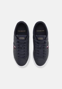 Pár námořnických modrých tenisek Tommy Hilfiger s bílými podrážkami, červenými a bílými bočními pruhy a tkaničkami, z pohledu shora na bílém pozadí.