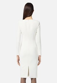 Frau mit langen, dunklen Haaren, die ein figurbetontes, langärmliges, knielanges weißes Kleid trägt und mit dem Rücken zu einer einfarbigen Hintergrund steht.