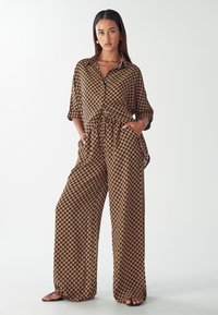 Smeđi kombinezon s uzorkom, ležernog kroja, s kragnom, kratkim rukavima i širokim hlačama. Čini se da je materijal lagan i prozračan.