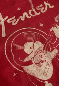 Personnage de vinyle souriant jouant de la guitare électrique avec le texte "Fender" sur un fond rouge texturé.