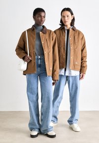 To personer har brune bomuldsjakker med trykknapper og brune kraveaccentuer, parret med løse blå denimjeans og hvide sko.