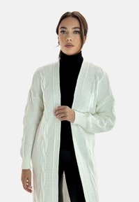 Cardigan blanco de punto trenzado con mangas largas y abertura frontal, con textura acanalada y detalles a lo largo de los bordes. Lleva encima un cuello de tortuga negro.