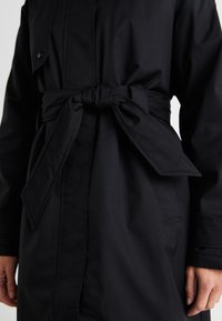 Abrigo negro de trench con cinturón en la cintura, que presenta una textura suave, botones en la parte delantera y bolsillos laterales. La tela parece duradera y resistente al agua.