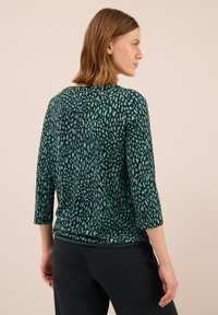 Frau mit schulterlangem Haar, die ein schwarzes und grünes gemustertes Langarmoberteil und schwarze Hosen trägt, seitlich vor einem schlichten Hintergrund stehend.