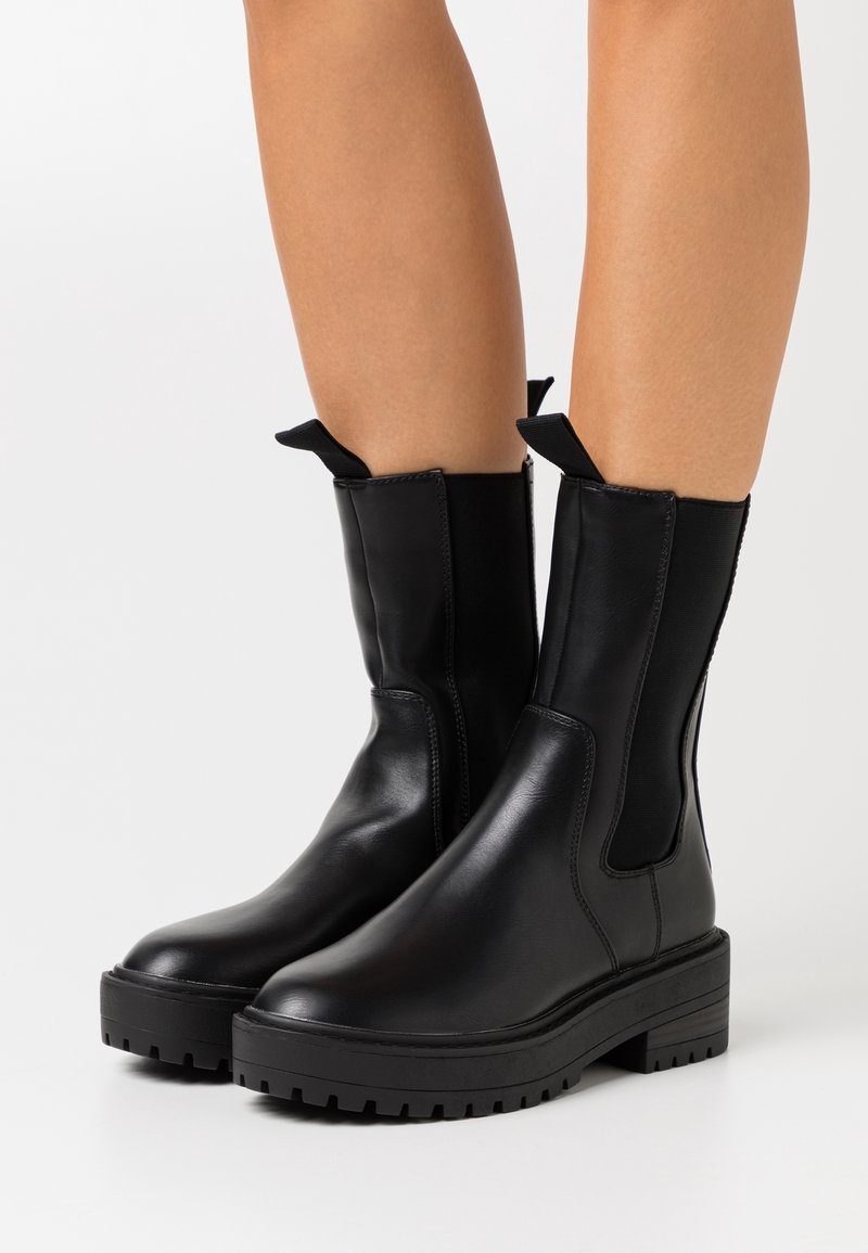 Bottes en cuir noir mi-mollet avec semelles épaisses et languettes, portées sur des jambes nues sur un fond neutre.