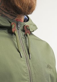 Veste à capuche verte avec fermeture éclair frontale, embouts en cordon marron et tissu texturé. Comprend une fermeture éclair argentée et un détail logo sur la poitrine.