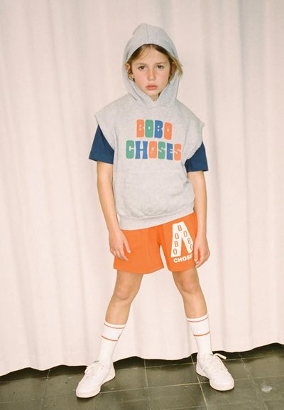 Šedá bezrukávová mikina s farebným nápisom "BOBO CHOSES", modré tričko s krátkym rukávom, oranžové šortky s logom, biele ponožky s červenými pruhmi a biele tenisky.