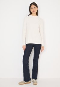 Pull-over en maille beige clair avec un col montant et une texture côtelée, associé à un jean évasé foncé et des baskets beiges avec des accents blancs.