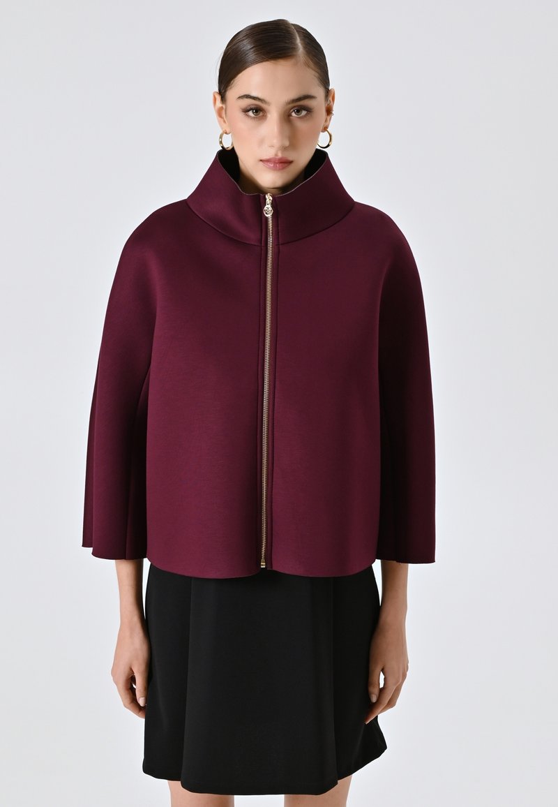 Bordeauxförmiger Reißverschluss-Cape mit hohem Kragen, aus glattem, strukturiertem Stoff gestaltet. Mit einem goldenen Reißverschluss ausgestattet, über einem schwarzen Kleid getragen.