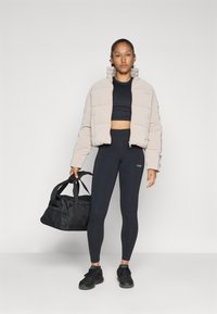 Lys beige polstret jakke med høy krage, svart crop-top, høyt liv svarte leggings og en svart treningsbag, mot en hvit bakgrunn.