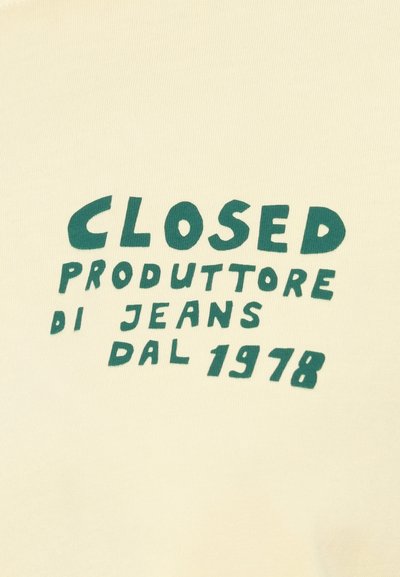 Tissu couleur crème avec texte vert : « CLOSED PRODUTTORE DI JEANS DAL 1978. » Le texte est en gras, au design ludique ; texture lisse.