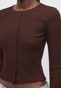 Gros plan d'une personne portant un haut marron côtelé ajusté à manches longues avec boutons, un col rond et un ourlet festonné à la taille.
