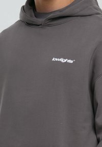 Sudadera gris hecha de una tela suave, con capucha ajustable y un pequeño logo blanco en el pecho. Diseño simple sin patrones visibles.