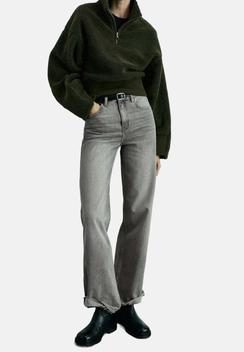 Personne portant un pull duveteux vert foncé, un jean gris clair taille haute et retroussé, une ceinture noire et des bottines noires, debout avec une main dans la poche.