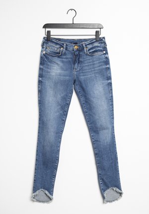 Modré vybledlé úzké džíny s třepenými lemy zavěšené na černém klipovém ramínku na bílém pozadí.