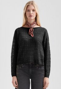 Blonde vrouw in een zwart kanten gebreide trui, donkere spijkerbroek met metalen oogjes, en een gebonden bruine patroon sjaal om de nek, staande tegen een witte achtergrond.