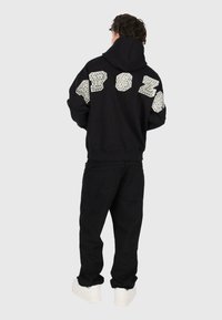 Mann in schwarzem Hoodie mit großen weißen gemusterten Buchstaben auf dem Rücken, schwarzen Hosen und weißen Sneakern, der von hinten steht.