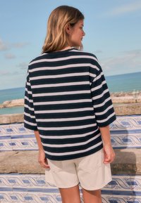 Navy and white striped oversized shirt with three-quarter sleeves, paired with light-colored shorts. Smooth fabric, relaxed fit.