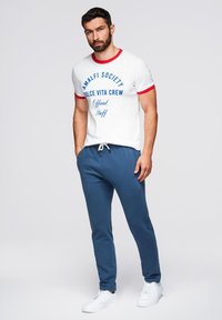 Vit kortärmad t-shirt med röd kant och blå text, kombinerad med marinblå sweatpants och vita sneakers. Enkel design, avslappnad stil.