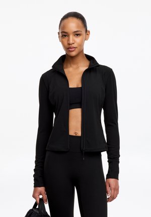 Femme portant une veste de sport noire, un soutien-gorge de sport et des leggings, tenant un sac de sport noir sur fond blanc.