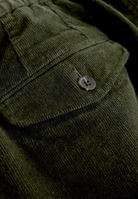 Dark green corduroy fabric featuring a pocket with a rounded flap and a four-hole button. Textured ribbed surface adds depth.