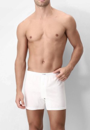 Uomo muscoloso con boxer bianchi, che sistema la cintura con la mano destra, su sfondo chiaro uniforme.