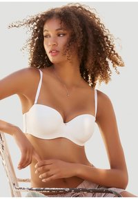 Sieviete ar cirtainiem matiem, nēsājot baltu strapless krūšturi, sēž uz rūpnieciskā metāla krēsla un skatās malā pret vienkrāsainu fonu.