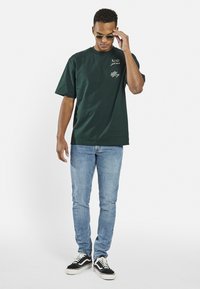 Grüne Baumwoll-T-Shirt mit kurzen Ärmeln, Logo-Detail auf der Vorderseite, kombiniert mit hellblauen Skinny-Jeans und schwarz-weißen Sneakern.