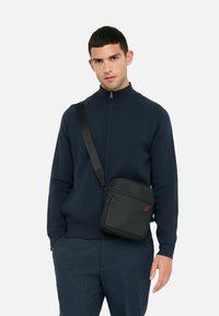 Mies, joka pelaa sinistä vetoketjullista takkia ja sinisiä housuja, ja jolla on musta olkalaukku roikkumassa olkapäältä, taustalla on pelkkä valkoinen tausta.