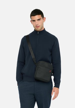 Mężczyzna w granatowej kurtce na zamek i niebieskich spodniach, z czarną torbą na ramię zawieszoną na ramieniu, na tle jednolitego białego tła.