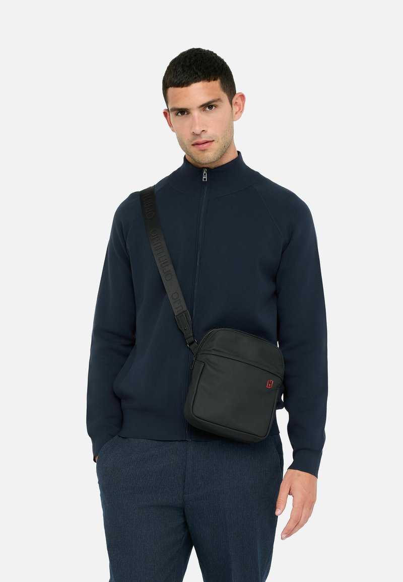 Mies, joka pelaa sinistä vetoketjullista takkia ja sinisiä housuja, ja jolla on musta olkalaukku roikkumassa olkapäältä, taustalla on pelkkä valkoinen tausta.