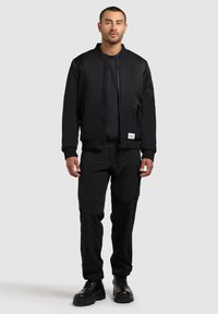 Giacca bomber nera con chiusura a zip frontale, polsini a costine e piccola etichetta. Abbinate a pantaloni cargo neri e stivali neri con lacci.